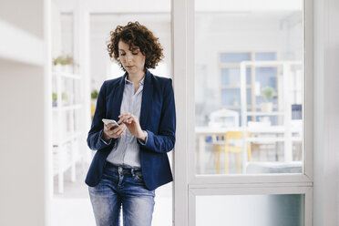 Businesswoman standing in office door, using smartphone - KNSF01578