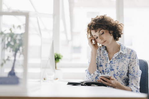 Geschäftsfrau im Büro mit Smartphone und Terminkalender, besorgt dreinblickend, lizenzfreies Stockfoto