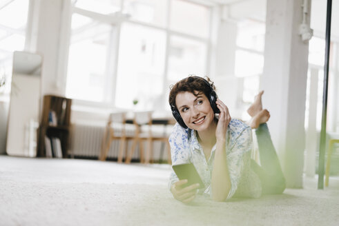 Woman lying on floor in her apartment, using smartphone and headphones - KNSF01596