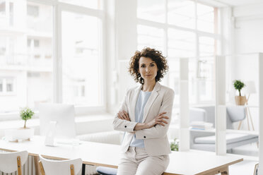 Businesswoman in office sitting on desk with arms crossed - KNSF01600