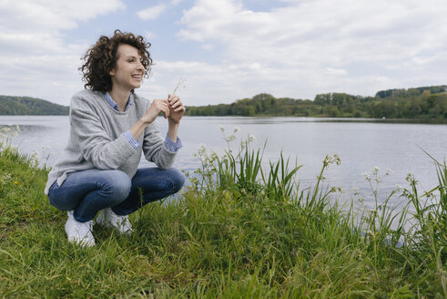 Frau hockt am Seeufer und riecht an einem Grashalm - KNSF01619