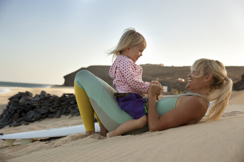 Spanien, Fuerteventura, glückliche Mutter und Tochter am Strand neben dem Surfbrett bei Sonnenuntergang - MFRF00873