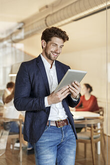 Lächelnder Geschäftsmann, der ein Tablet im Büro benutzt, mit einer Besprechung im Hintergrund - PESF00649