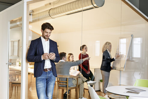 Geschäftsmann, der im Büro ein Mobiltelefon benutzt, mit einer Besprechung im Hintergrund - PESF00656