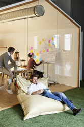 Man in office using tablet in bean bag with meeting in background - PESF00675