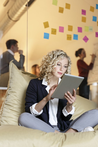 Frau im Büro mit Tablet in Sitzsack mit Sitzung im Hintergrund, lizenzfreies Stockfoto