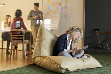 Woman in office using tablet in bean bag with meeting in background - PESF00683