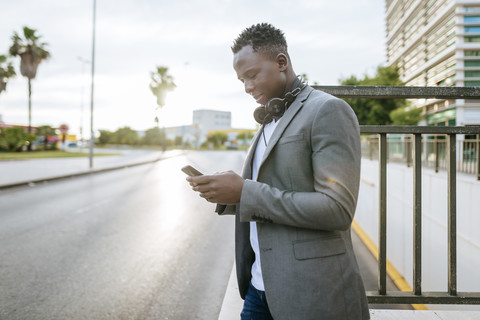 Junger Mann steht am Straßenrand und schaut auf sein Smartphone, lizenzfreies Stockfoto