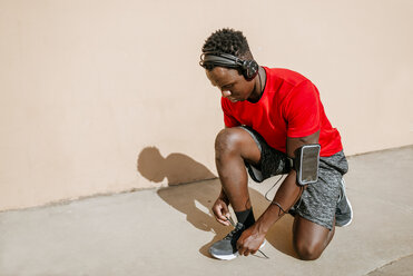 Man in sportswear tying his shoes while listening music with headphones - KIJF01574