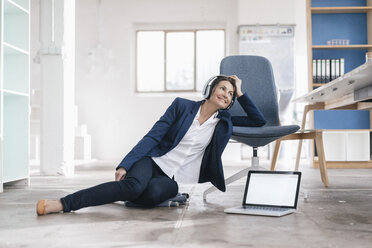 Smiling businesswoman sitting on the floor in a loft listening music with headphones - JOSF01167