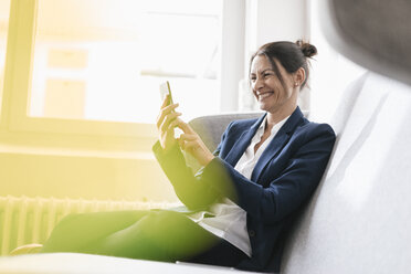 Laughing businesswoman sitting on a couch using her cell phone - JOSF01169