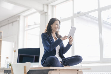 Businesswoman sitting on desk in a loft using tablet - JOSF01185