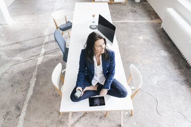 Pensive businesswoman sitting on desk in a loft with electronic devices - JOSF01188