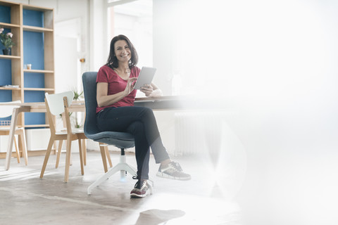 Porträt einer lächelnden Frau mit Tablet, die an einem Schreibtisch in einem Loft sitzt, lizenzfreies Stockfoto