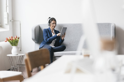 Geschäftsfrau sitzt auf einer Couch in einem Loft und benutzt Tablet und Kopfhörer, lizenzfreies Stockfoto