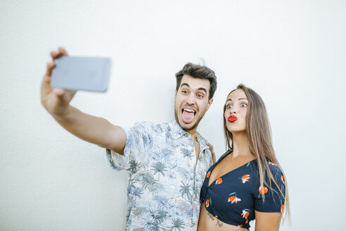 Verspieltes Paar macht ein Selfie mit Smartphone vor einer weißen Wand - KIJF01626