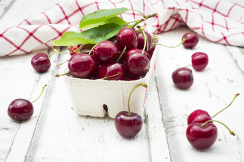 Cardboard box of cherries, leaves and kitchen towel on white wood stock photo