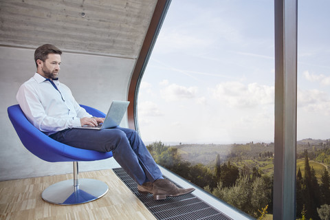 Geschäftsmann sitzt auf einem Stuhl in einem Dachbodenbüro und benutzt einen Laptop, lizenzfreies Stockfoto