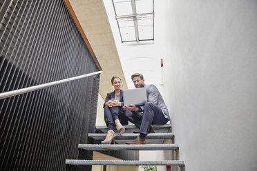 Geschäftsmann und Geschäftsfrau sitzen auf einer Treppe im Büro und teilen sich einen Laptop - RHF01960