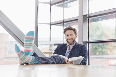 Portrait of smiling businessman putting his feet on the table reading book - RHF02001