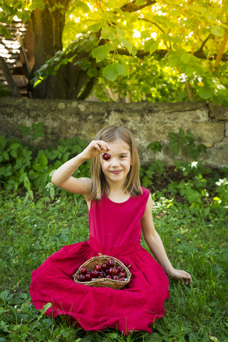 Porträt eines kleinen Mädchens im roten Sommerkleid auf einer Wiese sitzend mit einem Korb voller Kirschen, lizenzfreies Stockfoto