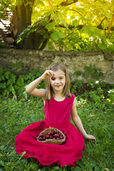 Portrait of little girl wearing red summer dress sitting on a meadow with basket of cherries - LVF06180