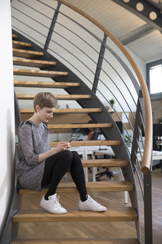 Frau sitzt auf einer Treppe in einem modernen Büro und schaut auf ihr Handy, lizenzfreies Stockfoto