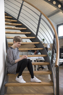 Frau sitzt auf einer Treppe in einem modernen Büro und schaut auf ihr Handy - FKF02340