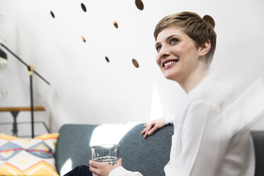 Smiling woman holding glass of water on couch - FKF02341