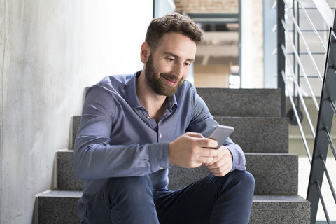 Lächelnder Mann sitzt auf einer Treppe und schaut auf sein Handy, lizenzfreies Stockfoto
