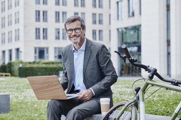 Portrait of smiling mature businessman outdoors with laptop, takeaway coffee and bicycle - RORF00872