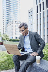 Happy mature businessman outdoors with laptop and takeaway coffee - RORF00883