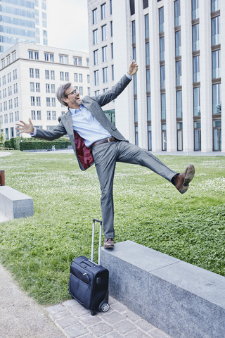 Playful mature businessman with rolling suitcase balancing on wall taking a selfie stock photo