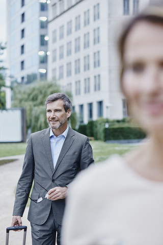 Lächelnde Geschäftsfrau im Freien mit Laptop und Geschäftsmann im Hintergrund, lizenzfreies Stockfoto