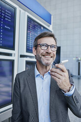 Lächelnder Geschäftsmann mit Handy am Fahrplan auf dem Flughafen - RORF00937