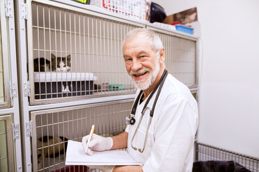 Lächelnde leitende Tierärztin beim Ausfüllen von Dokumenten in einem Käfig mit Katzen - HAPF01689