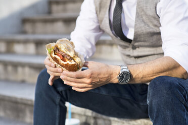 Businessman in the city sitting on stairs eating a hamburger - HAPF01722