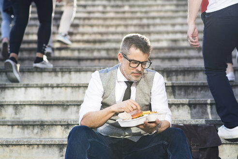 Businessman in the city sitting on stairs eating a hamburger - HAPF01723