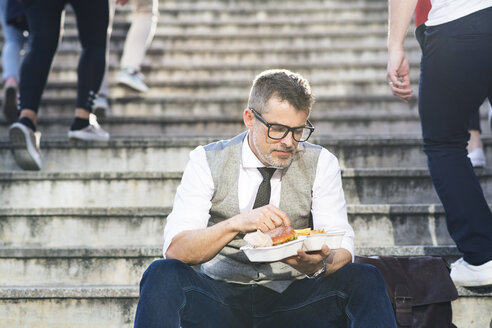 Geschäftsmann in der Stadt, der auf einer Treppe sitzt und einen Hamburger isst - HAPF01723