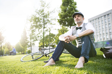 Mature businessman with bicycle in the city park sitting on grass - HAPF01728