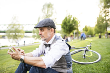 Confident mature businessman with bicycle and smartphone in the city park sitting on grass - HAPF01731