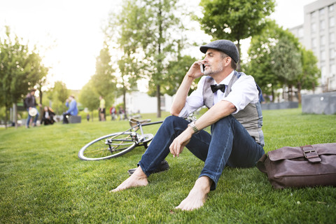 Selbstbewusster reifer Geschäftsmann mit Fahrrad und Smartphone im Stadtpark auf dem Rasen sitzend, lizenzfreies Stockfoto