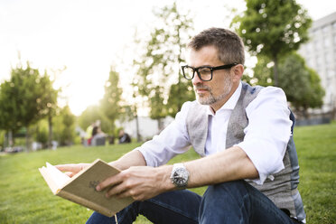 Confident mature businessman in the city park sitting on grass reading book - HAPF01739