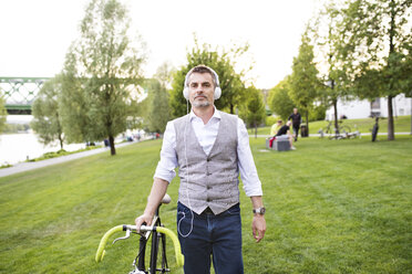 Selbstbewusster reifer Geschäftsmann mit Fahrrad und Kopfhörern im Stadtpark - HAPF01743