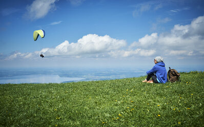 Germany, Chiemgau, hiker sitting on Alpine meadow watching paraglider - DIKF00255