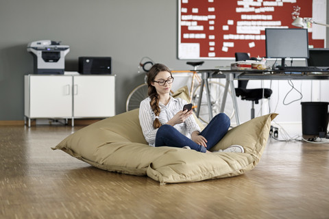 Young woman with cell phone sitting in bean bag in office stock photo