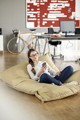 Young woman with cell phone sitting in bean bag in office - PESF00724