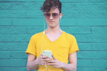 Portrait of young man listening music with smartphone and earphones in front of blue brick wall - RTBF00907