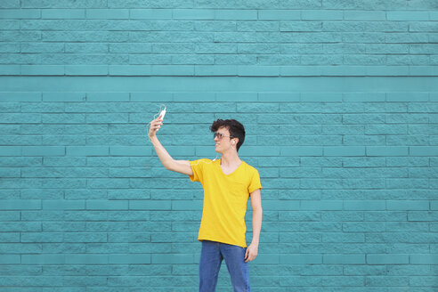 Junger Mann macht ein Selfie mit Smartphone vor einer blauen Backsteinmauer - RTBF00908