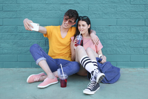 Junges Paar mit alkoholfreien Getränken macht Selfie mit Smartphone vor blauer Backsteinmauer - RTBF00915